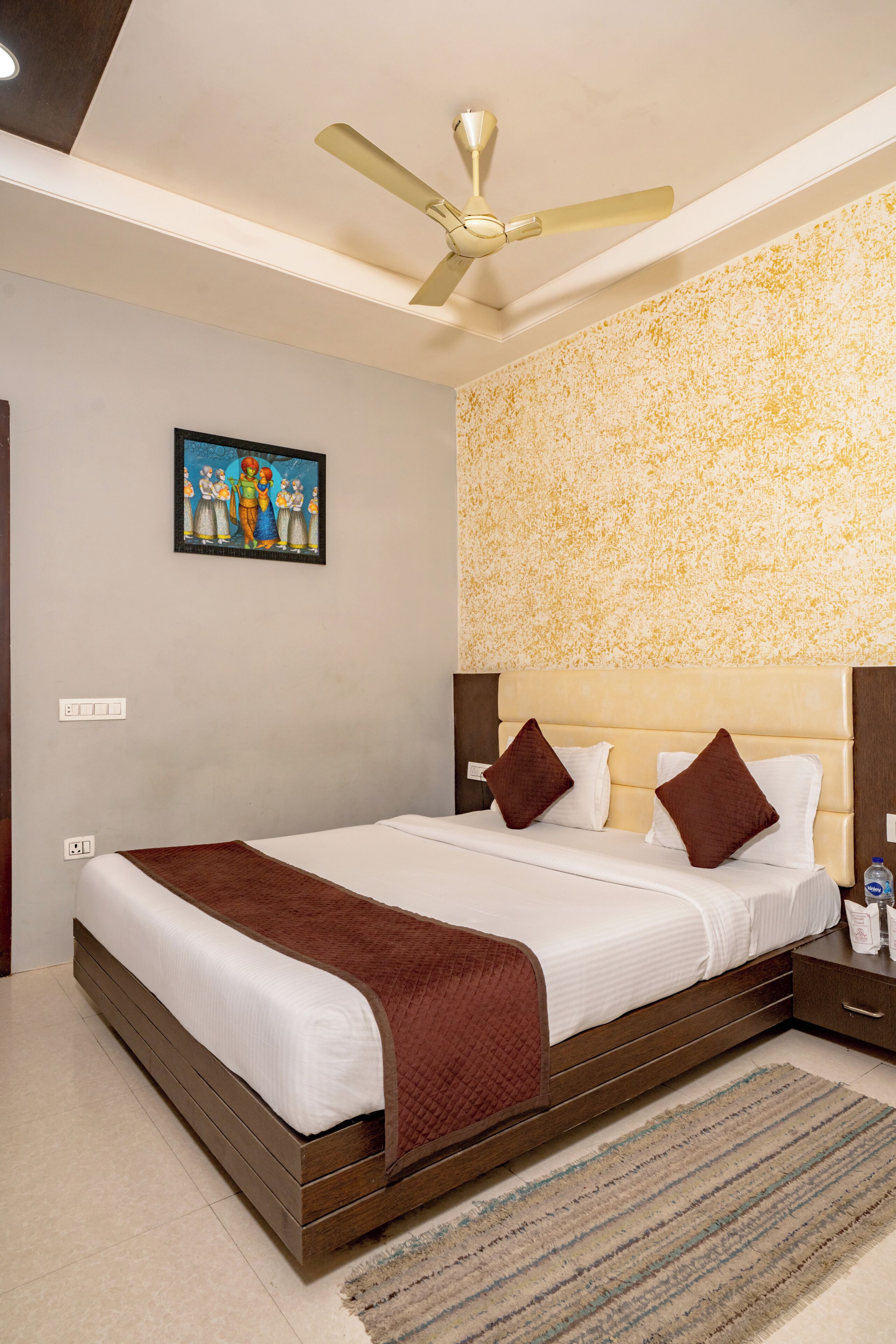 A neatly made bed with a brown throw blanket, a ceiling fan, and a painting on the wall.