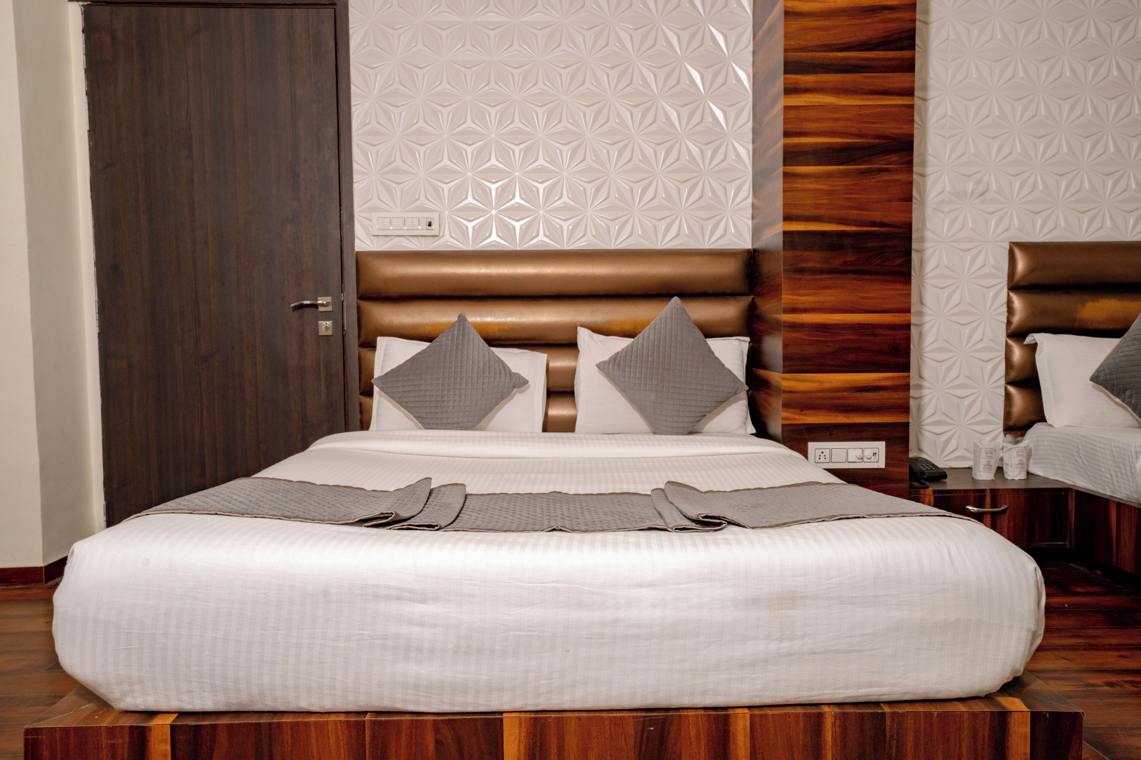 A neatly made bed with a headboard, a wooden floor, and a patterned wall.