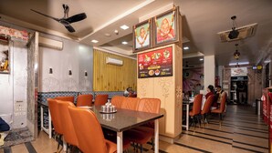 A restaurant with a dining area featuring a long table with orange chairs, a wall-mounted menu, and a ceiling fan. - Hotel Sawamani and Restaurant (Danta Ramgarh)