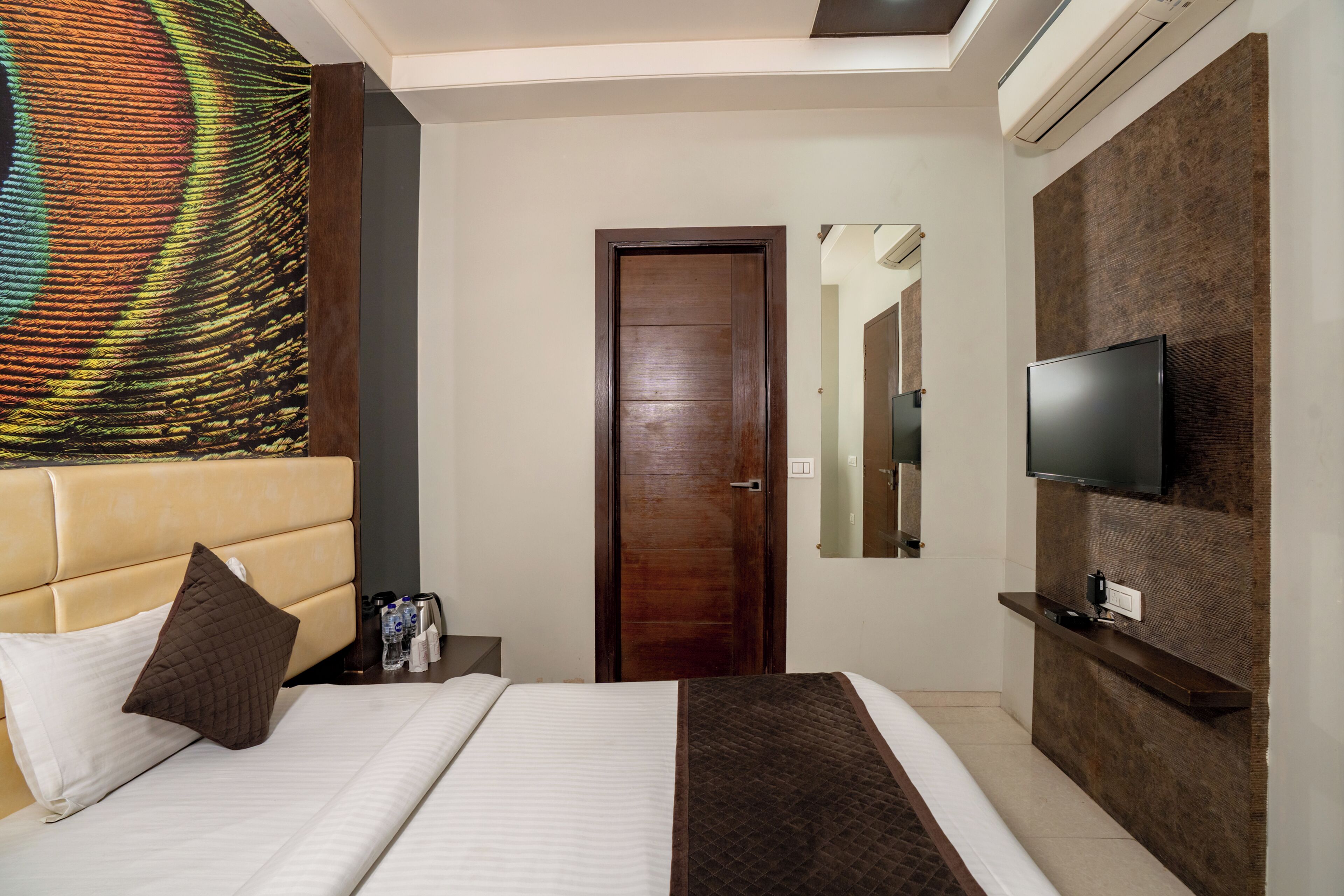 A hotel room with a bed, a TV, a wooden door, and a large artwork on the wall.
