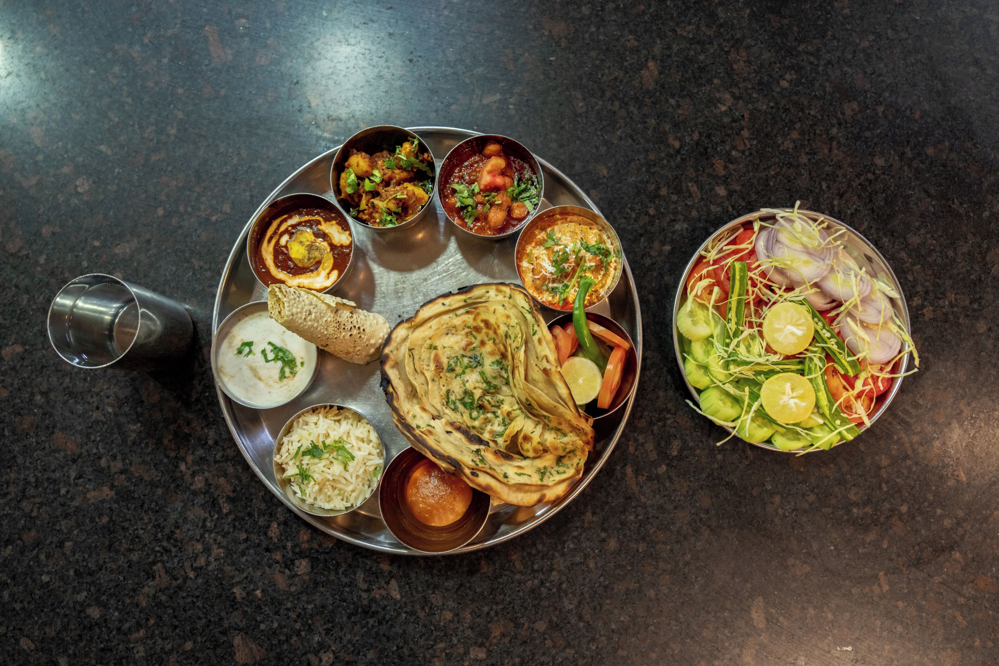 A meal with various dishes including flatbreads, curries, and a salad on a metal tray.