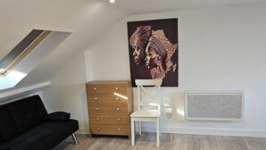 Una camera moderna con soffitto spiovente, un divano nero, una cassettiera in legno, una sedia bianca e un grande quadro astratto.