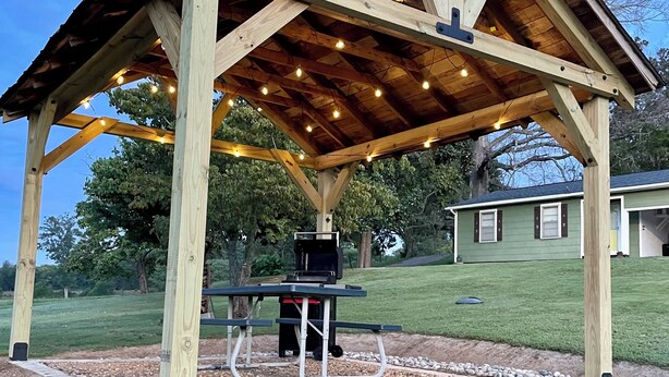Un belvédère en bois équipé d’un barbecue et d’un espace salon, entouré d’un chemin de gravier et de verdure.