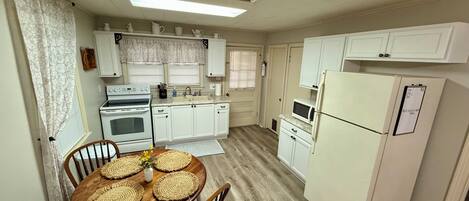 A compact kitchen with white cabinets, a refrigerator, a microwave, and a stove. There is a dining table with placemats and a vase of flowers.