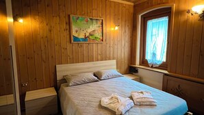 Un dormitorio con paneles de madera, una cama, almohadas, una mesita de noche, un cuadro y una ventana con cortinas.