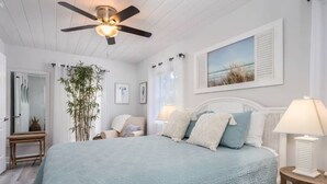 Un dormitorio con cama, ventilador de techo, mesita de noche, lámpara, ventana con contraventanas e imagen enmarcada en la pared.