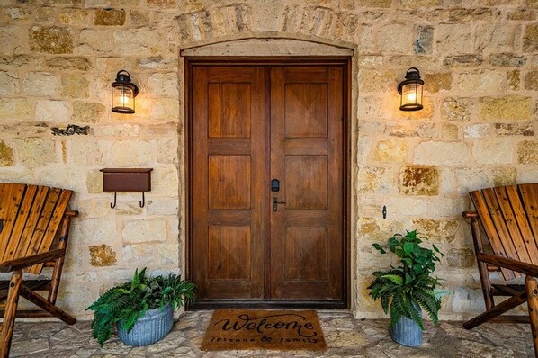 A stone wall entrance with a wooden double door, two wall-mounted lanterns, and two wooden chairs.