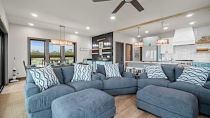 A modern living room with a sectional sofa, a ceiling fan, and a kitchen area in the background. - Magnolia by the Sea: Beachfront Home, Private Pool, Mother-In-Law, Secluded Beac (Indian Pass)