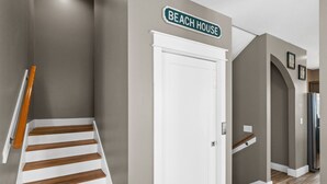 A hallway with a staircase, a white door, and a sign that reads "BEACH HOUSE".