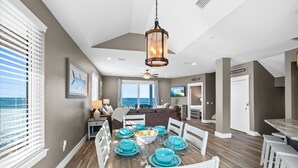 A dining area with a wooden table set for six, white chairs, and a view of the ocean through large windows.