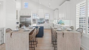A spacious kitchen with white cabinetry, a central island, and a dining area with wicker chairs. - Estate Home: Gulf Views, Private Pool, Pool Table, Sleeps 20! (Mexico Beach)