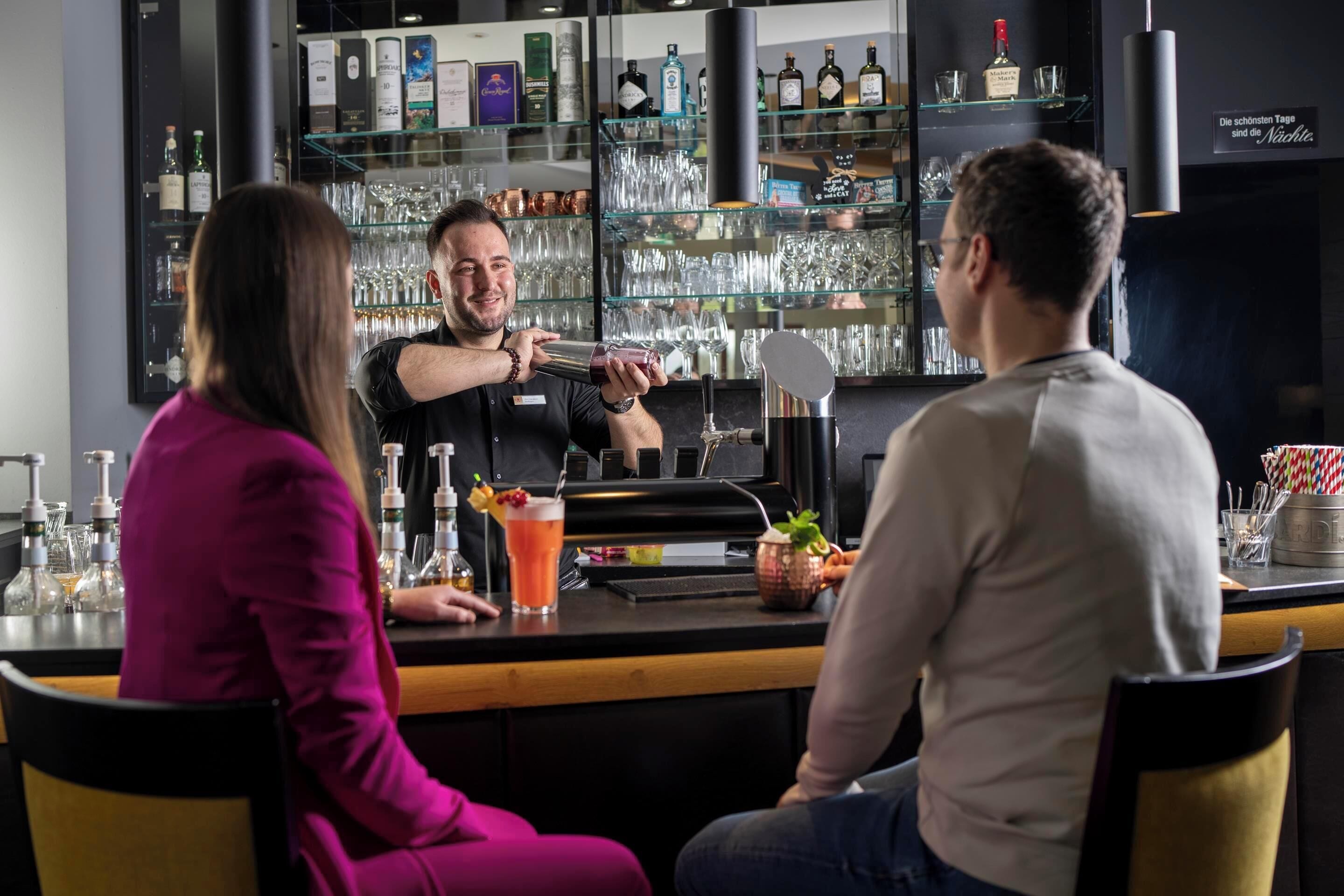 A bar area with a bartender preparing drinks, shelves with bottles and glasses, and a counter with a menu.
