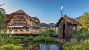 A traditional chalet with a balcony, a pond, and a wooden building with a sign. - Mosers Blume Relax & Genuss Hotel (Haslach im Kinzigtal)