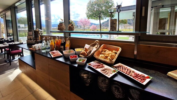 A breakfast buffet with various food items and beverages, a coffee machine, and a view of a landscape with trees and a body of water through large windows. - Mosers Blume Relax & Genuss Hotel (Haslach im Kinzigtal)