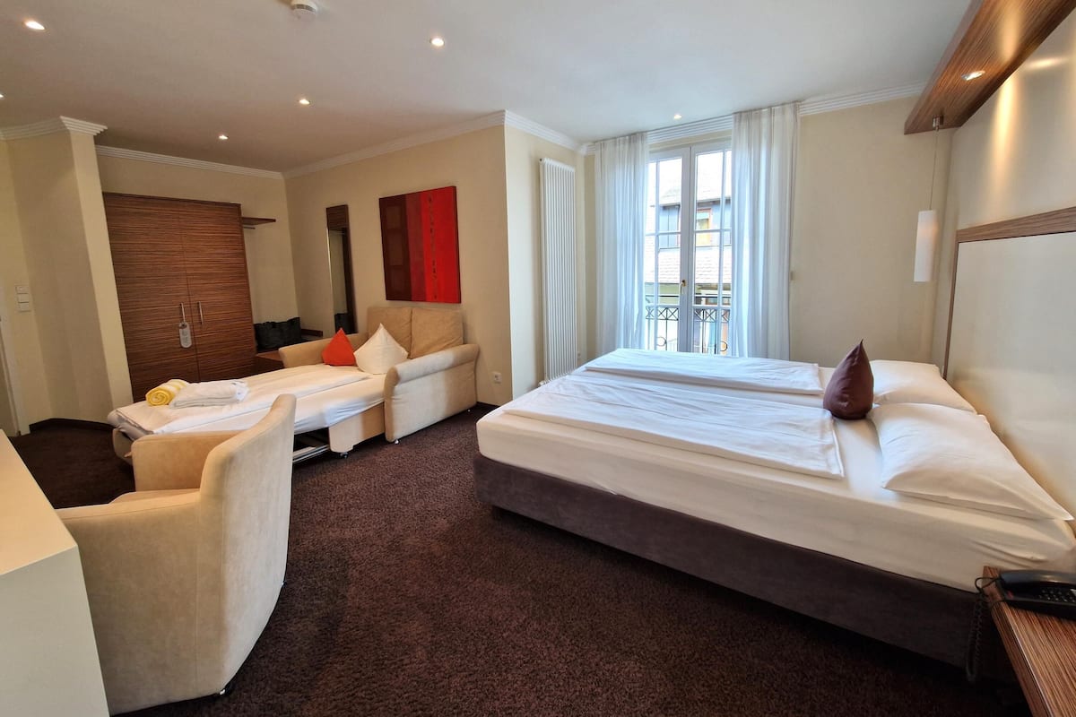 Ein Hotelzimmer mit einem Bett, zwei Sesseln, einer Couch, einem Holzschrank, einer roten Akzentwand und einem Fenster mit Blick nach draußen.