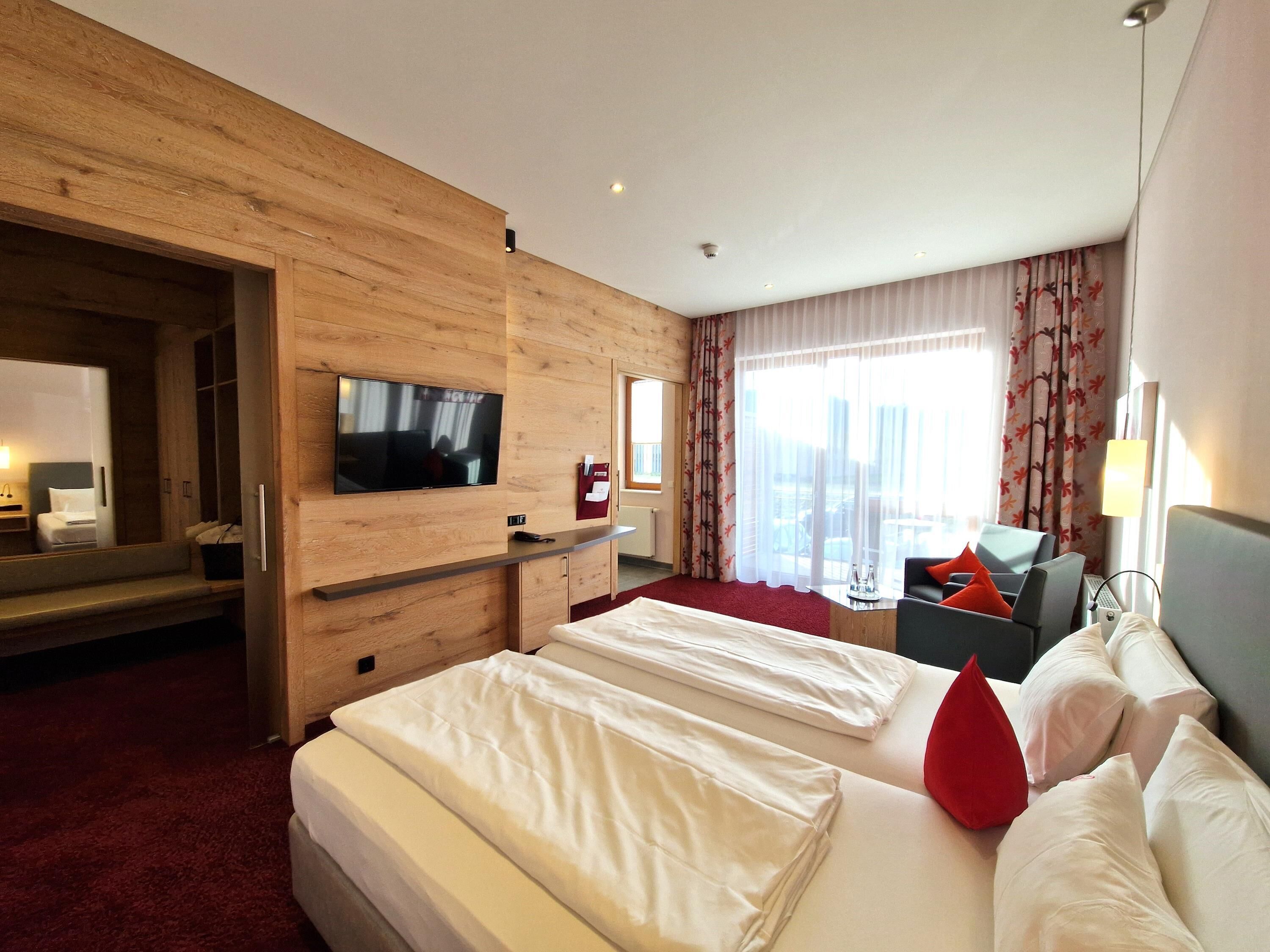 A hotel room with a wooden wall, a flat-screen TV, a bed with white linens, a sofa with red cushions, and a window with sheer curtains.