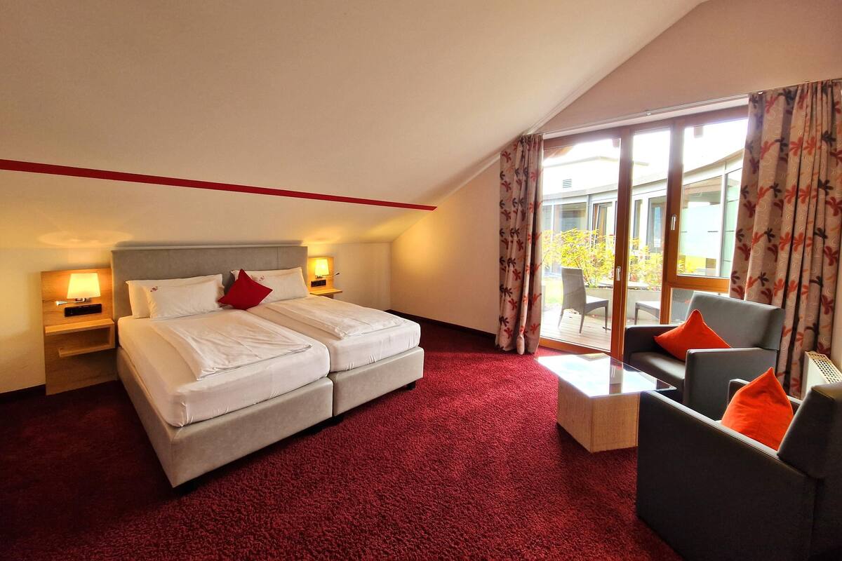 Ein Hotelzimmer mit einem großen Bett, zwei Nachttischen mit Lampen, einem kleinen Sofa und Blick ins Freie durch eine Schiebetür.