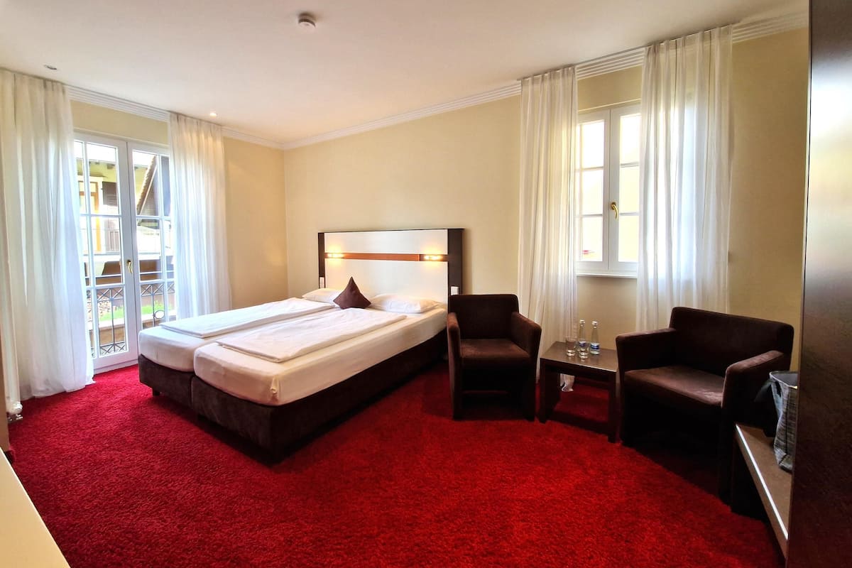 Ein Hotelzimmer mit einem großen Bett, zwei Sesseln, einem kleinen Tisch und einem Fenster mit Vorhängen.