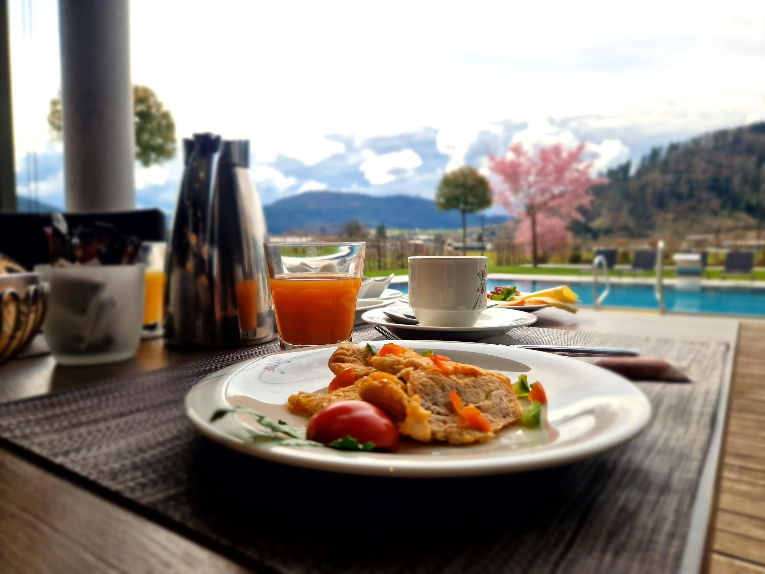 A breakfast setting with a plate of food, a glass of orange juice, and a cup, overlooking a pool and mountains.