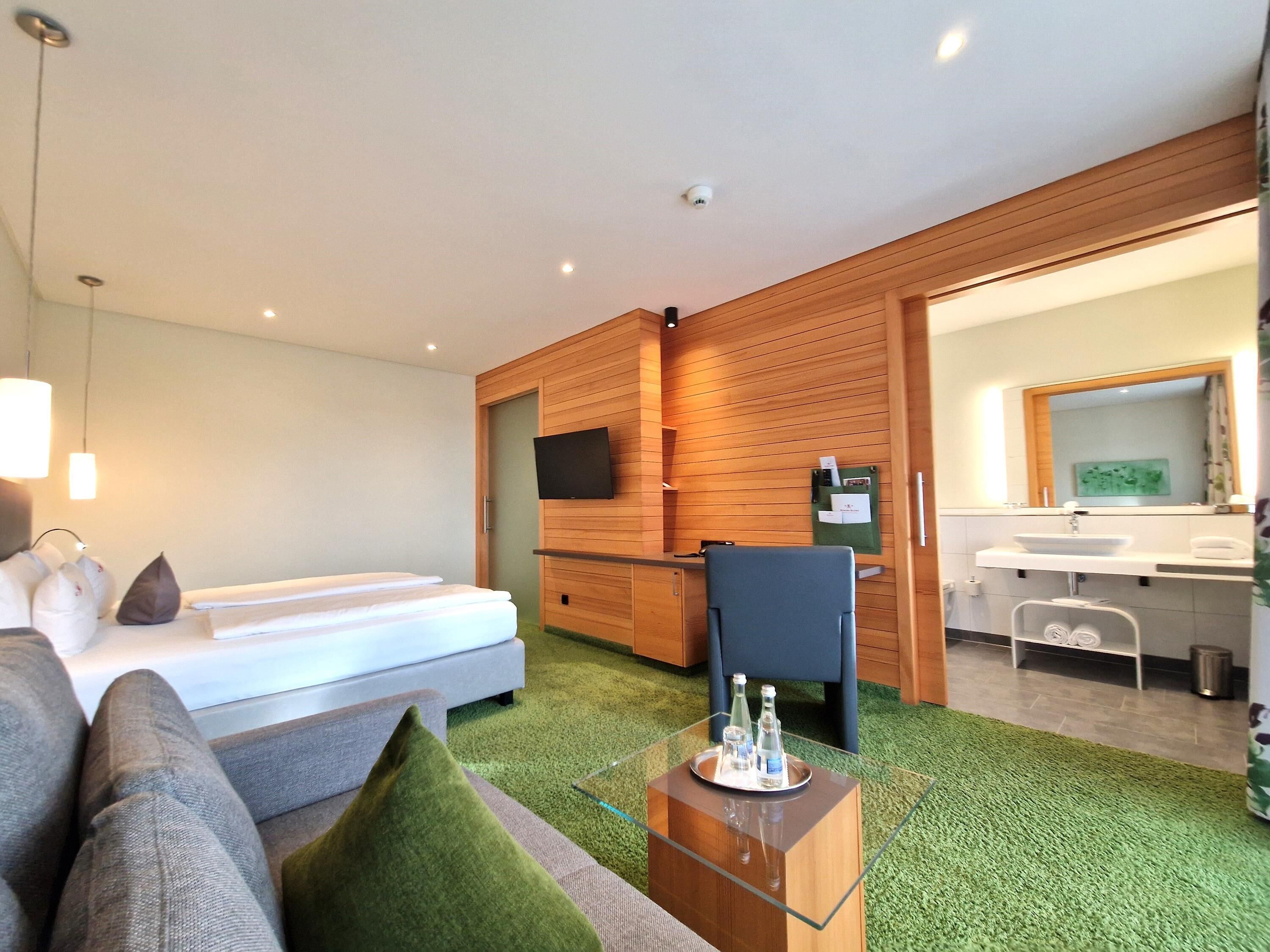 Een moderne hotelkamer met een bed, een zithoek met een glazen salontafel, een aan de muur bevestigde televisie, en een badkamer die door een open deur zichtbaar is.