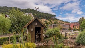A rustic wooden structure with a rooster weather vane, surrounded by greenery and a small pond. - Mosers Blume Relax & Genuss Hotel (Haslach im Kinzigtal)