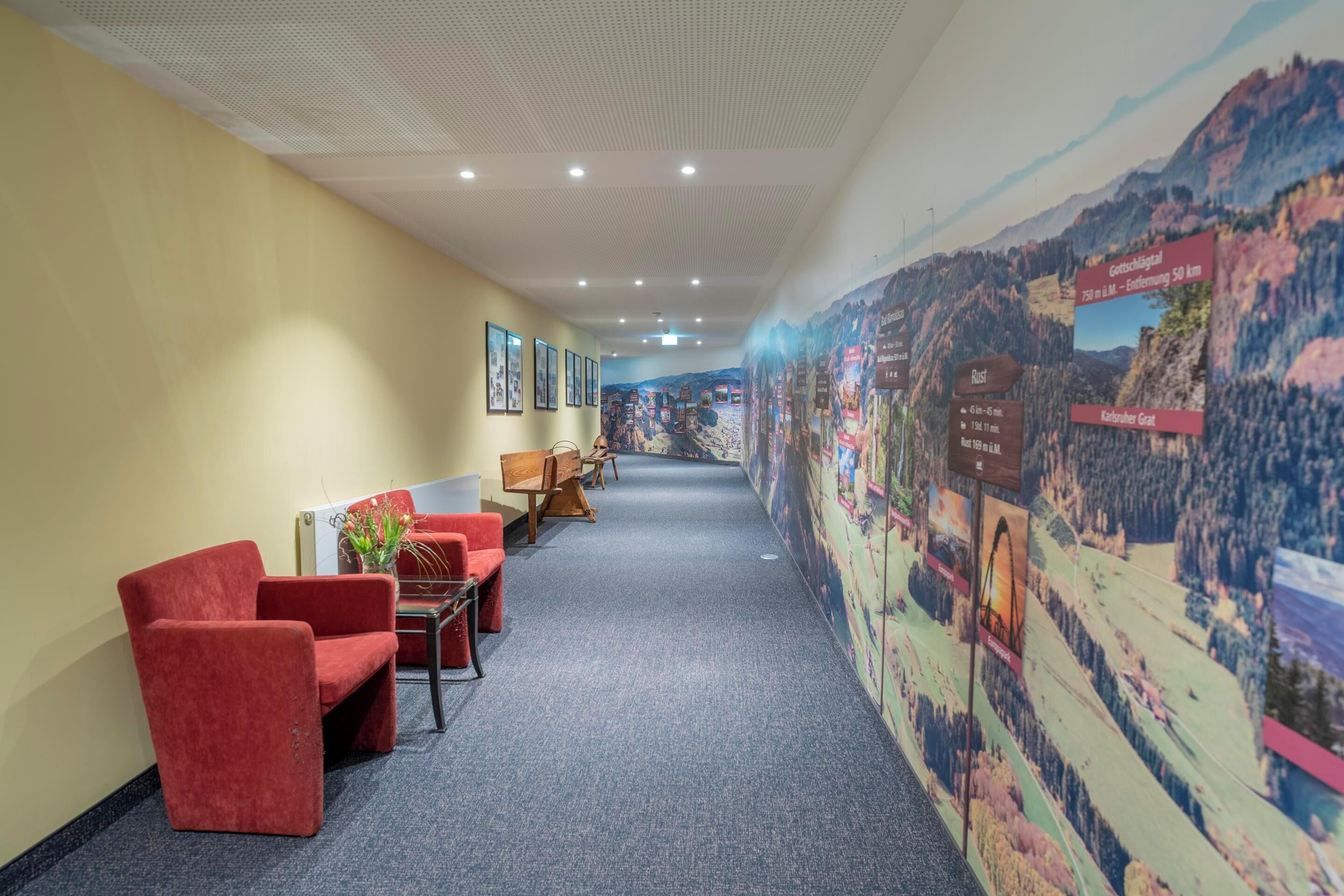 A hallway with red chairs, wooden tables, and a carpeted floor. There are framed pictures on the wall and a large mural depicting a mountainous landscape.