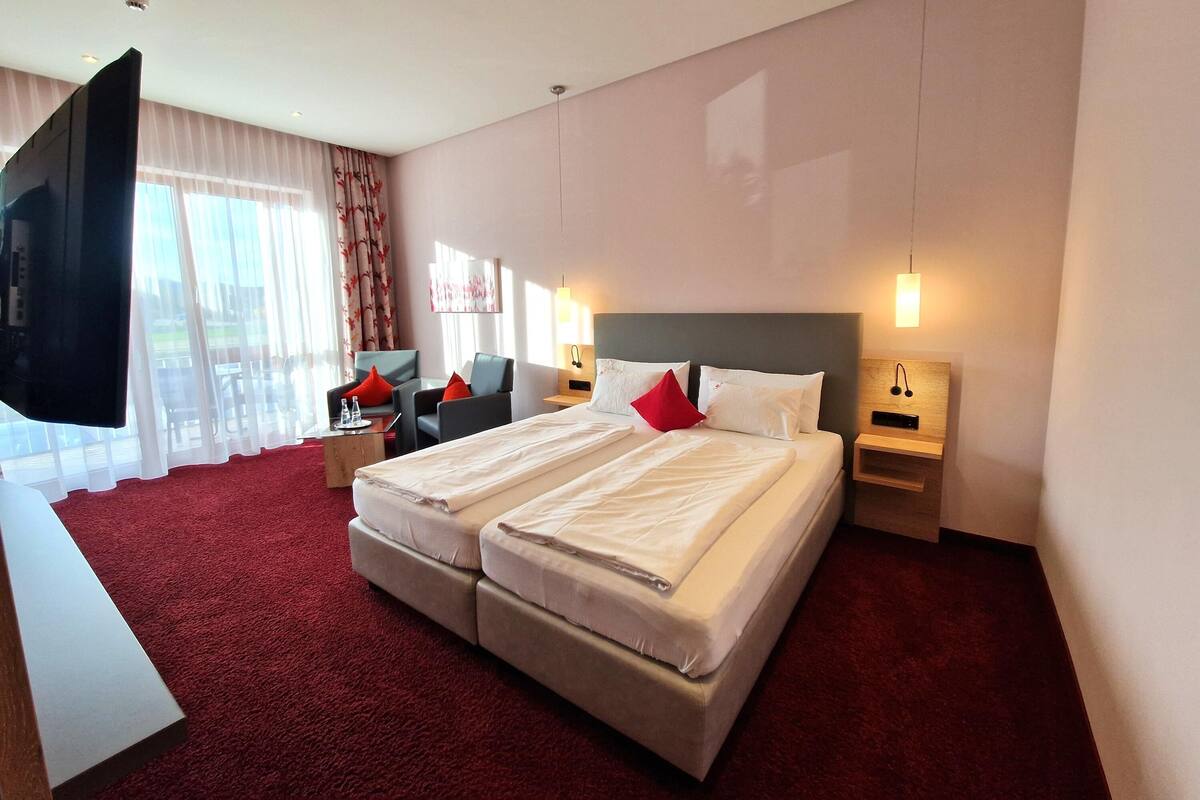 Ein Hotelzimmer mit einem großen Bett, zwei Nachttischen, einem Flachbildfernseher und Blick ins Freie durch leichte Vorhänge.
