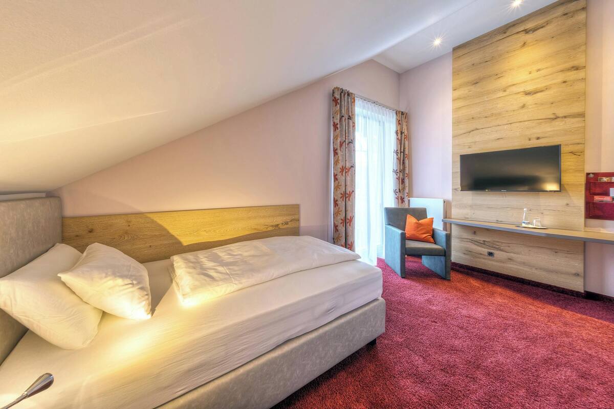 Ein modernes Hotelzimmer mit einem großen Bett, einem an einer Holzwand montierten Flachbildfernseher, einem Schreibtisch mit Stuhl und einem roten Teppich.