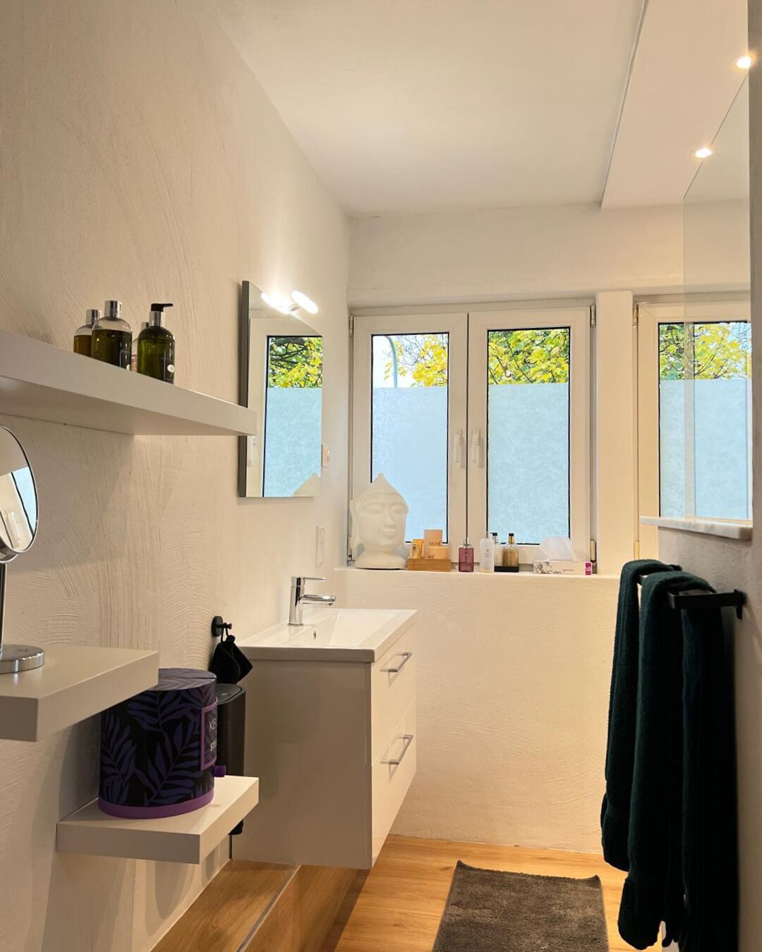 Une salle de bain moderne avec un lavabo blanc, un miroir et des étagères contenant des articles de toilette. Il y a des fenêtres donnant sur des arbres et un porte-serviettes avec une serviette.