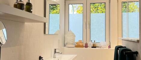 Une salle de bain moderne avec un lavabo blanc, un miroir et des étagères contenant des articles de toilette. Il y a des fenêtres donnant sur des arbres et un porte-serviettes avec une serviette.