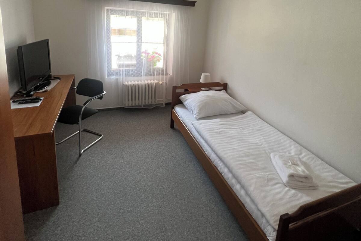 A small, clean room with a single bed, a desk with a computer, a chair, and a window with sheer curtains.