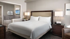 Une chambre d’hôtel avec un grand lit, une table de chevet avec une lampe, une commode et une salle de bain équipée d’un miroir et d’un porte-serviettes.