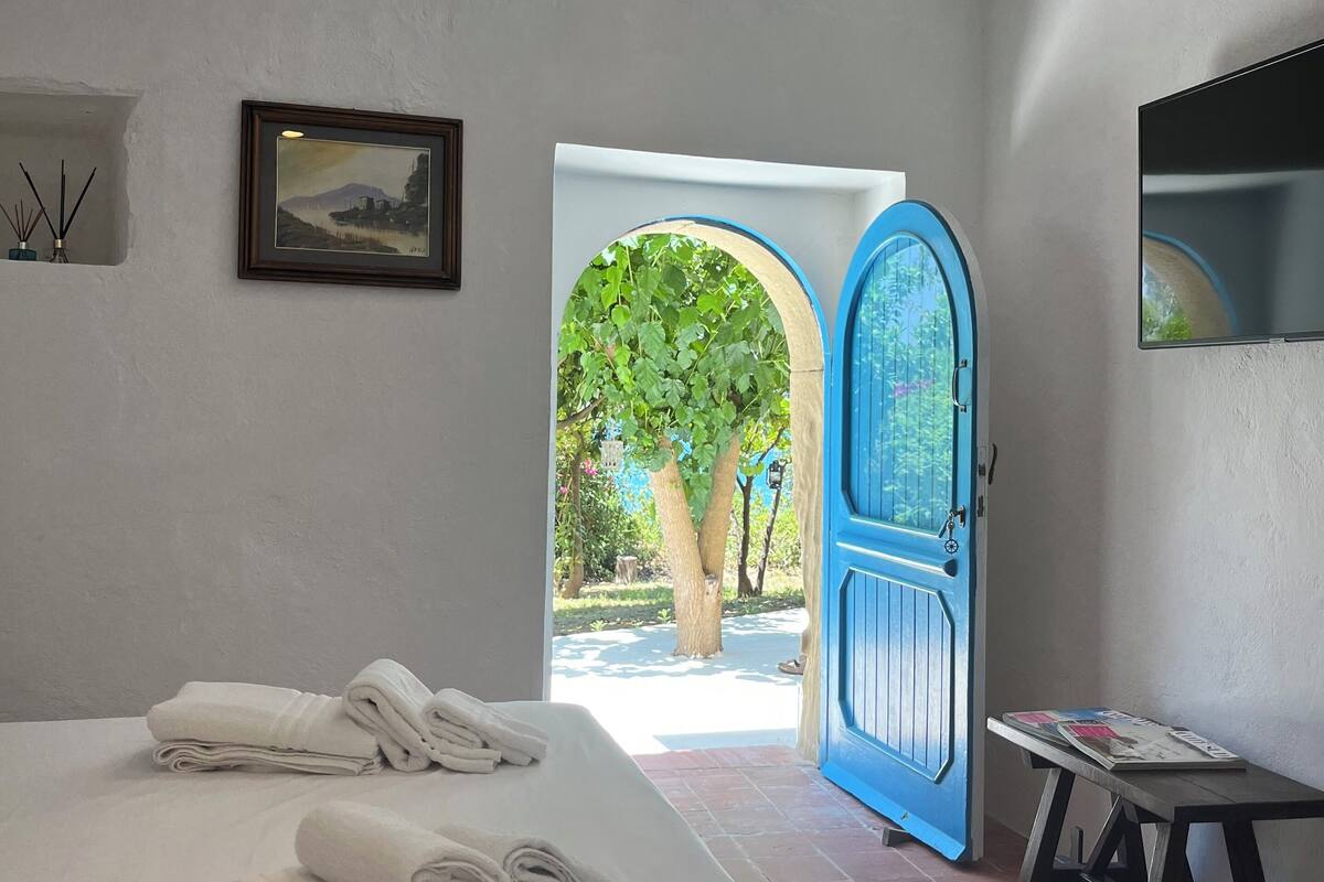 A bedroom with a bed, a framed picture, a blue door, and a small table with a magazine.
