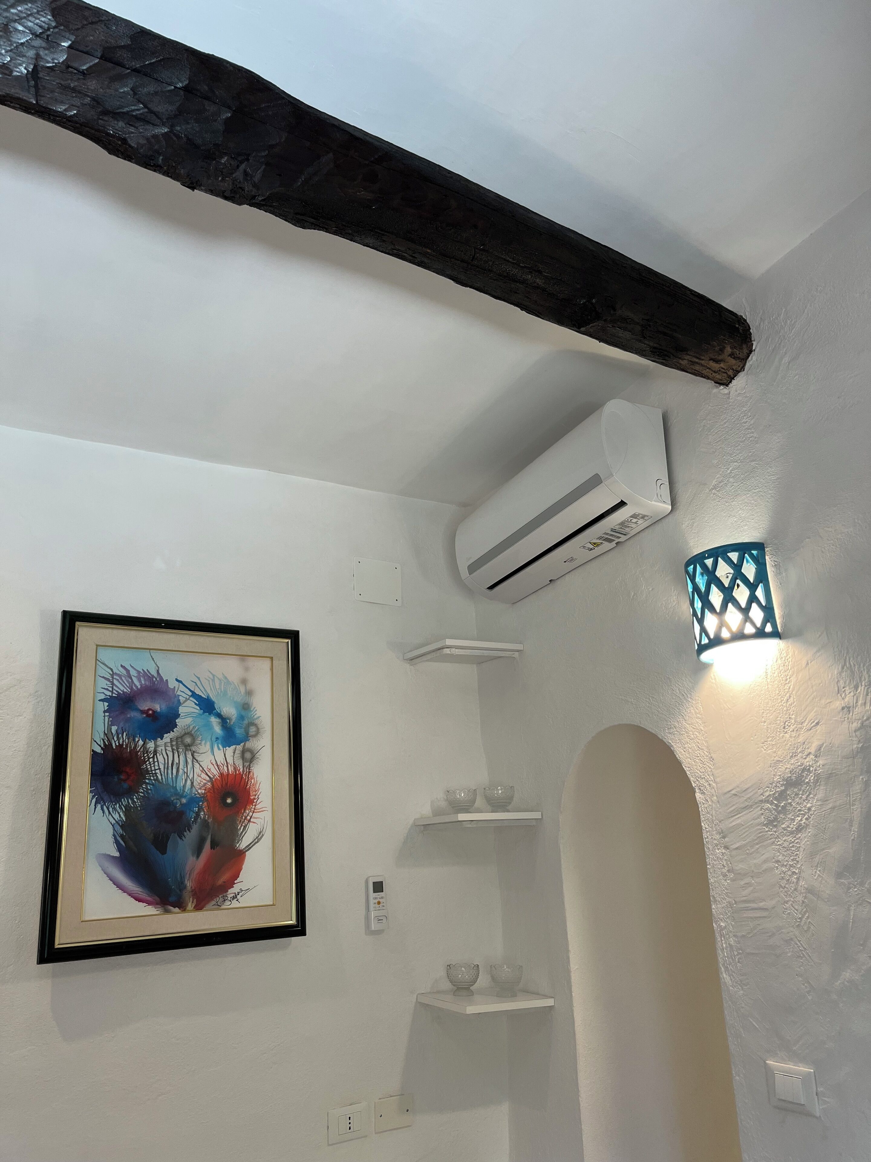 Ein Zimmer mit weißer Wand, einem gerahmten abstrakten Gemälde, einer weißen Klimaanlage, einer kleinen Ablage mit zwei Schalen und einer dekorativen Leuchte.