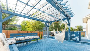Eine überdachte Terrasse im Freien mit einer Hängematte, einer Bank mit Kissen und einem Holzzaun im Hintergrund.