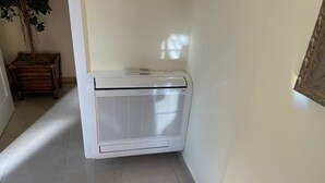 A white air conditioning unit in a corner of a room with tiled flooring and a wooden cabinet. - Enjoy KRK waterfalls, sun, beach and sea: Domicile Sunce Kornati (Skradin)