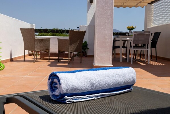 A sunlit terrace with a rolled towel on a chair, wicker furniture, and a table with chairs.