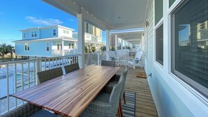 A covered outdoor area with a wooden table and chairs, a view of the ocean, and a building in the background. - Beach Retreat | Pool + Steps to Sand (Gulf Shores)