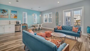 A modern living room with a blue sofa, a dining area with a wooden table, and a kitchen with white cabinets. - Beach Retreat | Pool + Steps to Sand (Gulf Shores)