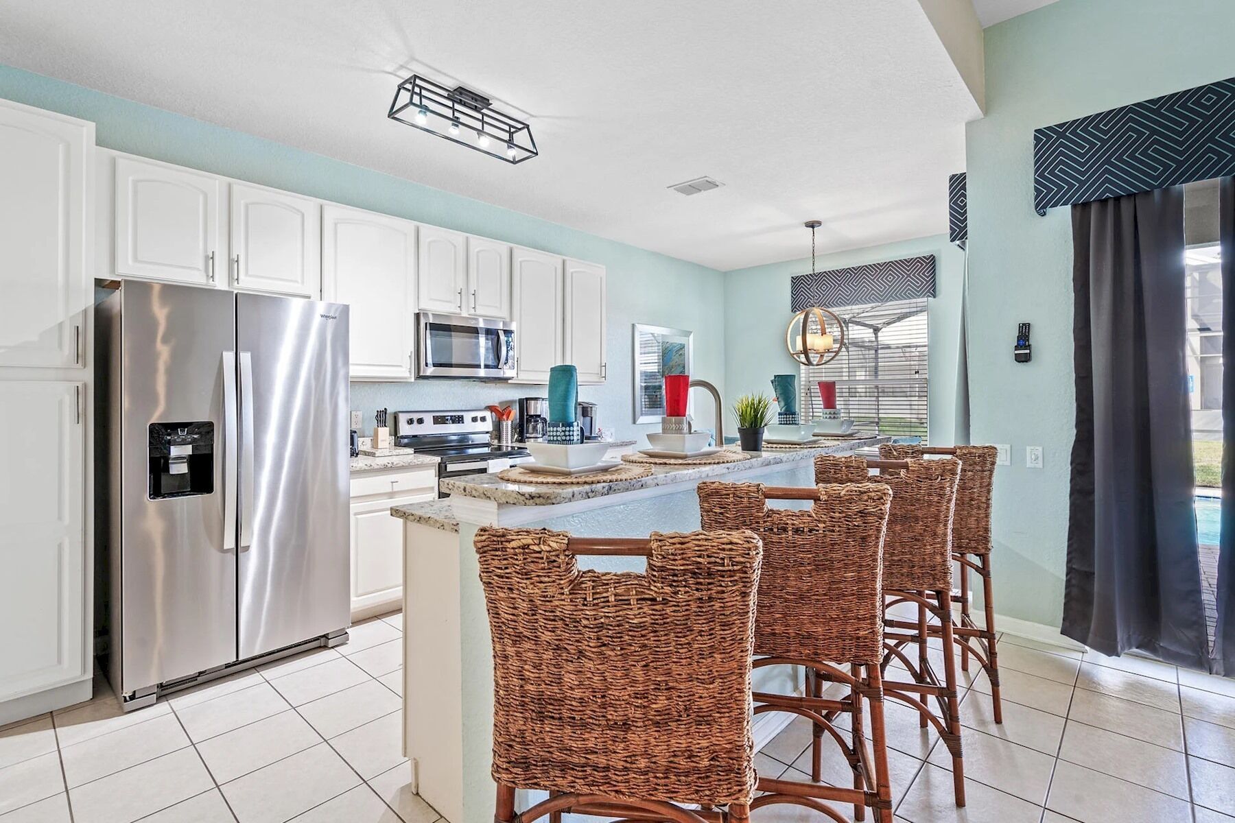 A modern kitchen with white cabinets, stainless steel appliances, a central island with bar stools, and a view of the outdoors through large windows.