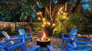 A fire pit with four blue Adirondack chairs arranged around it, surrounded by pebbles and tropical plants.