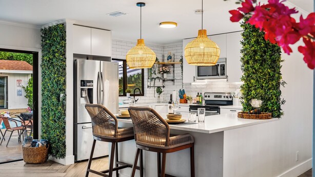 A modern kitchen with white cabinets, a central island with bar stools, hanging wicker pendant lights, and a vertical garden.