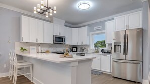 A modern kitchen with white cabinets, a stainless steel refrigerator, a microwave, and a large island with seating. - Modern Oceanview Vacation Home (Saint George Island)