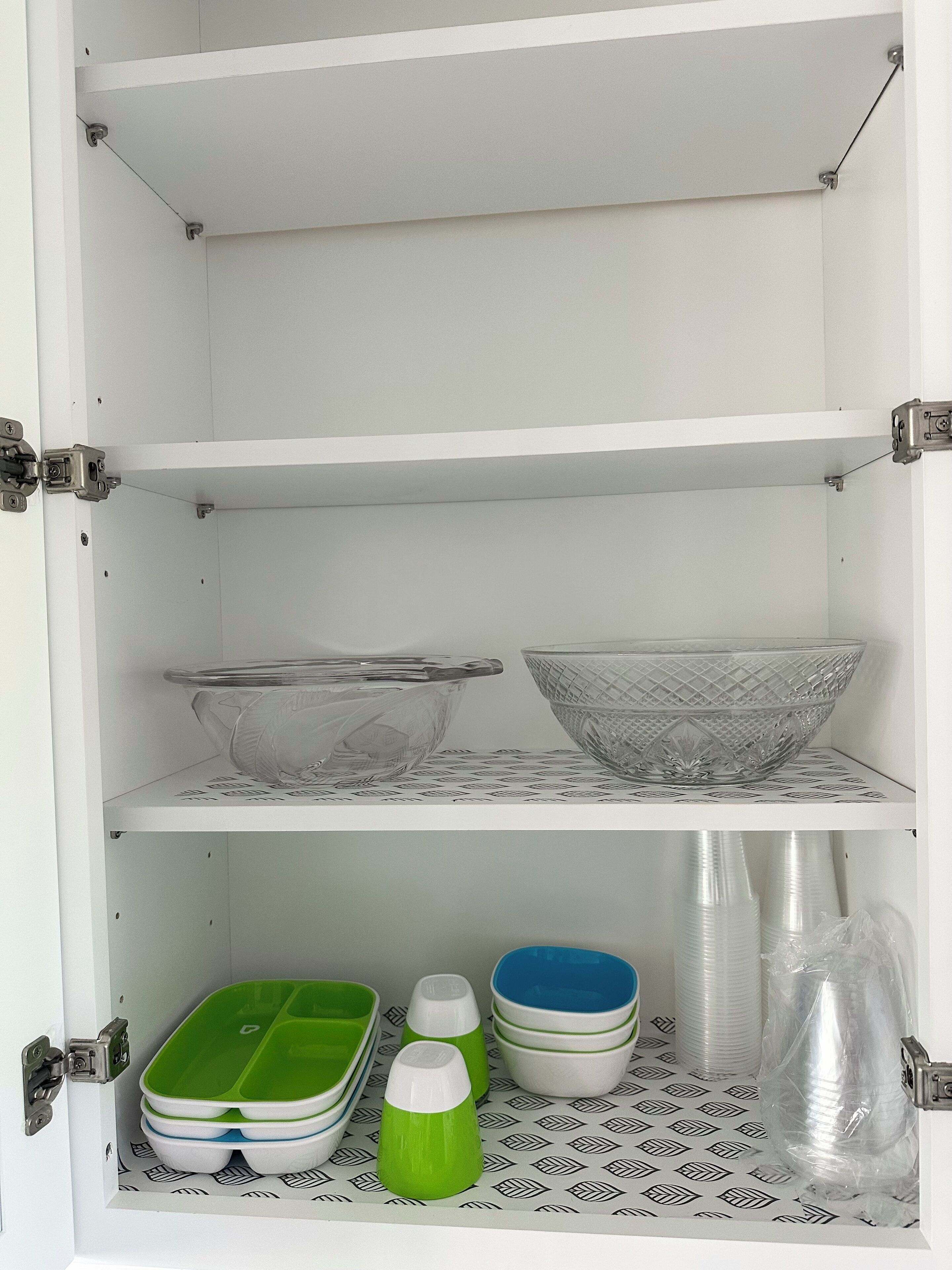 A white cabinet with two shelves; the top shelf holds two glass bowls, and the bottom shelf contains green and white containers, blue and white bowls, and plastic bottles.