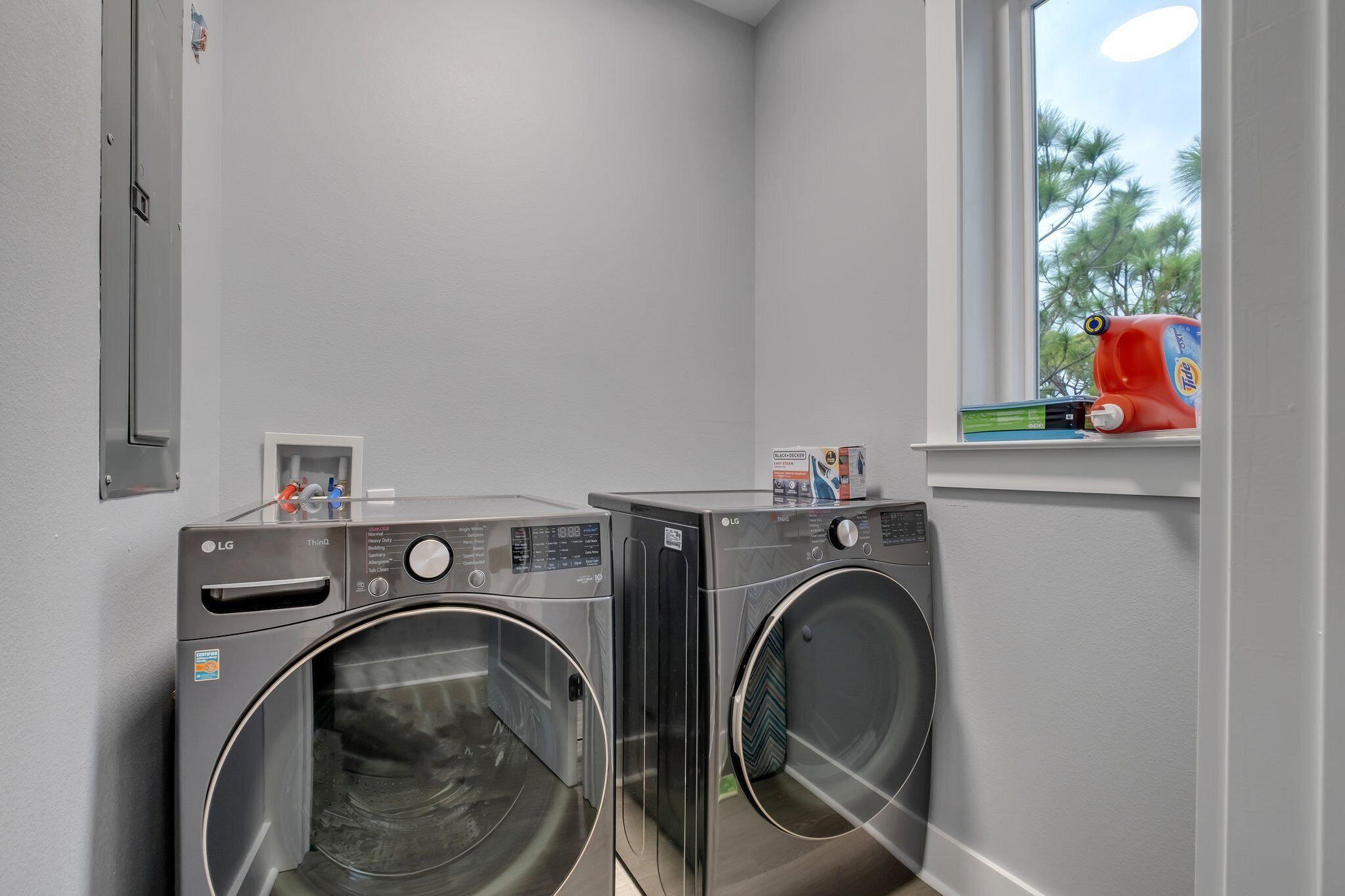 Une buanderie équipée de deux lave-linge et deux sèche-linge LG à chargement frontal, d’une fenêtre donnant sur les arbres et d’une étagère où sont rangées les produits de lessive.