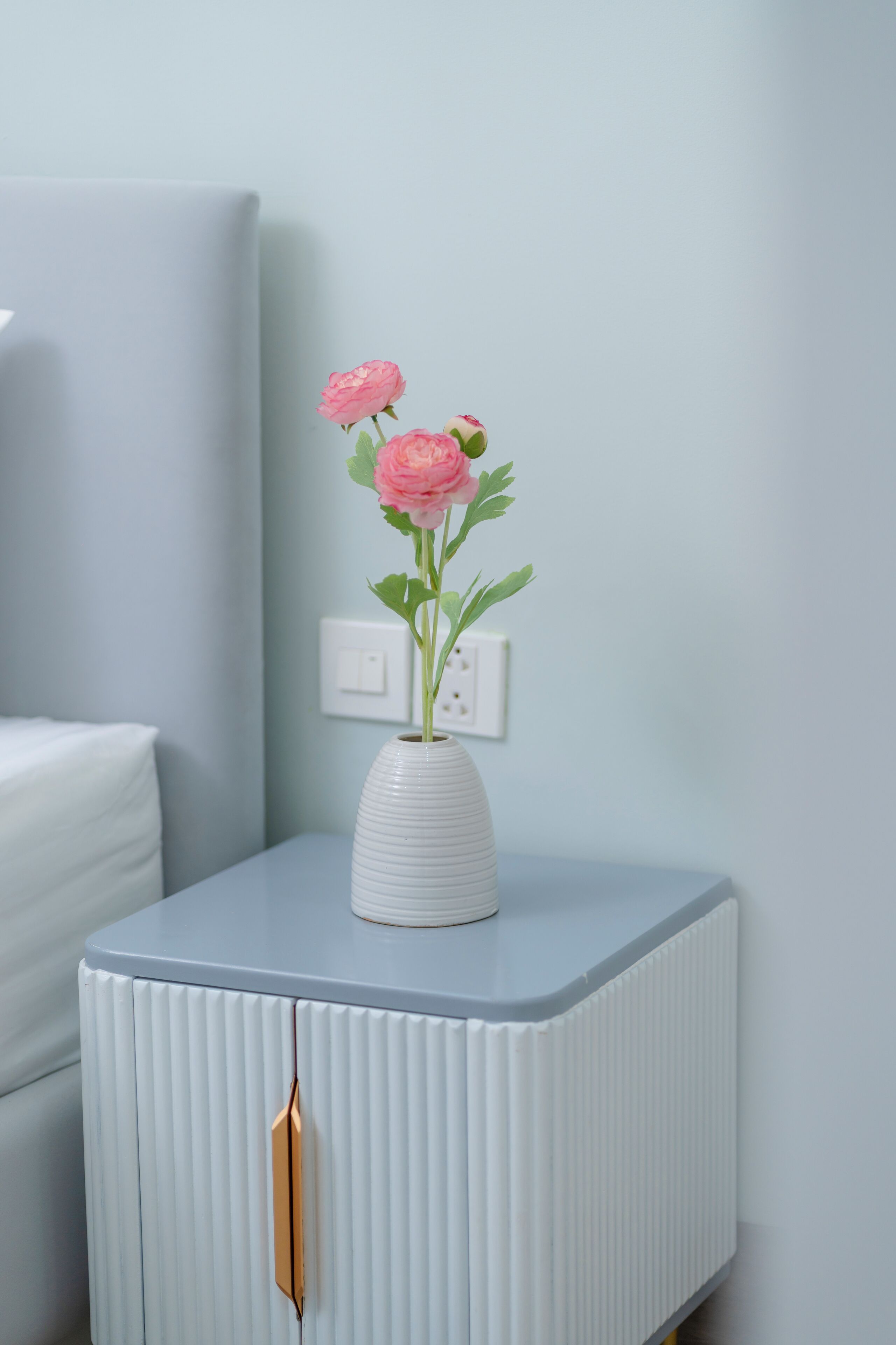 A white bedside table with a vase containing pink flowers, a white mattress, and a wall socket.