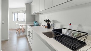Uma cozinha moderna com armários brancos, pia e bancada. Há uma janela com vista para árvores.