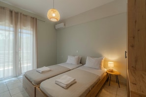 A hotel room with two beds, a window with curtains, a hanging light fixture, and a small table with a lamp.