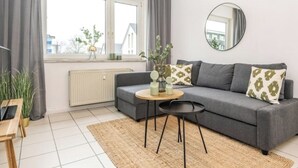 Ein modernes Wohnzimmer mit einem grauen Sofa, einem runden Spiegel, einem kleinen Holztisch und Blick ins Freie durch ein Fenster.