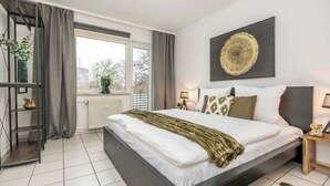 Ein modernes Schlafzimmer mit einem großen Bett, einem Fenster mit Vorhängen, einem Wandbild und einem Nachttisch mit Lampe.
